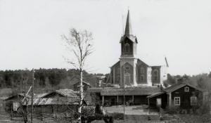 Mustavalkoinen valokuva 1900-luvun alusta. Maatilan rakennusten takana kohoaa kirkko.